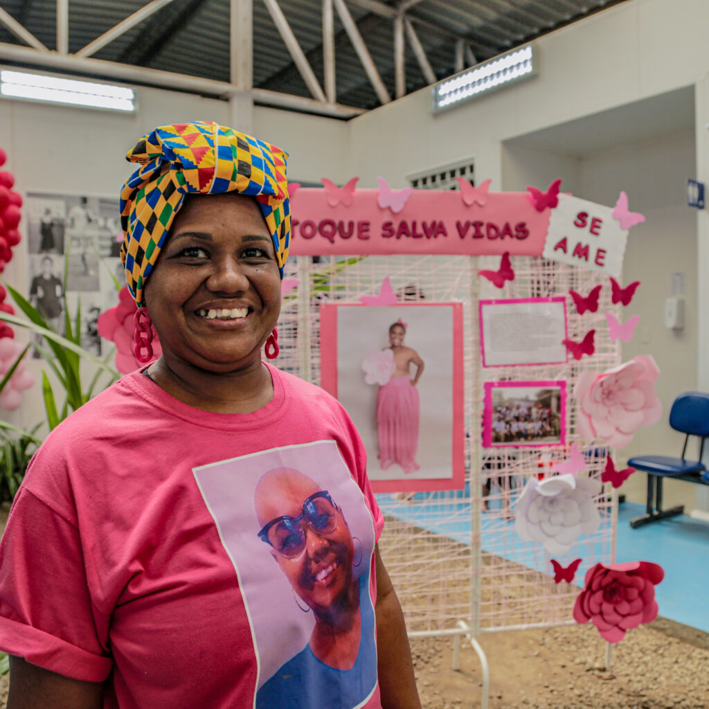 mulher sorri em frente a cartaz com sua foto e dizeres sobre o autoexame da mama